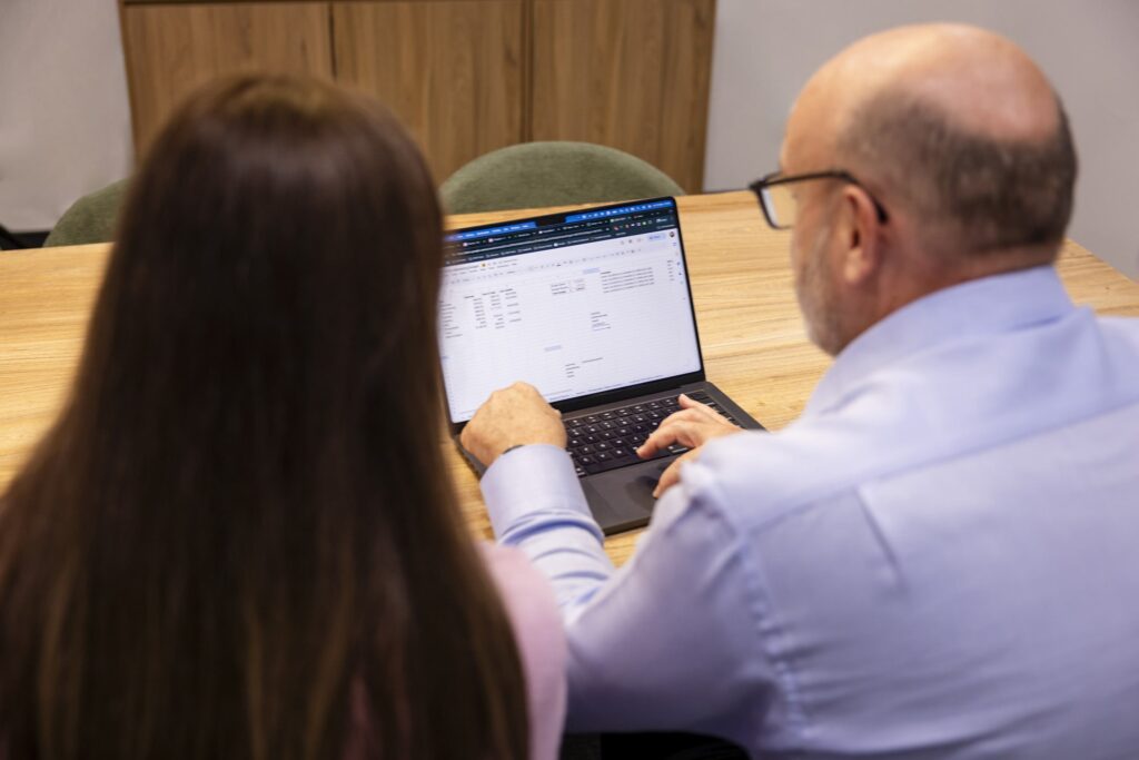 Month end spreadsheets: Two people viewed from behind at a wooden table, working on a laptop showing a spreadsheet